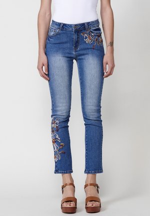 Jeans de mezclilla azul bordados con diseños florales en colores como rojo y blanco, con un corte ajustado y una longitud cropped, complementados por una cintura con botones.