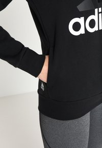Schwarzes Sweatshirt mit großem weißen Logo, ausgestattet mit einer Kängurutasche und gerippten Bündchen. Tragen über grauen Leggings. Weicher, strukturiertes Material.