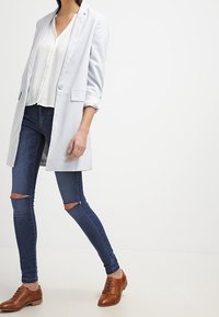 Hellblauer Blazer über einer weißen Bluse, kombiniert mit dunkelblauen Skinny-Jeans mit Knie-Rissen und braunen Leder-Schnürschuhen.