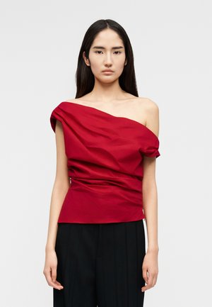 Γυναίκα με μακριά σκουρόχρωμα μαλλιά που φοράει κόκκινη ασύμμετρη μπλούζα με έναν ώμο έξω και μαύρο παντελόνι, στέκεται μπροστά σε απλό ανοιχτό φόντο.