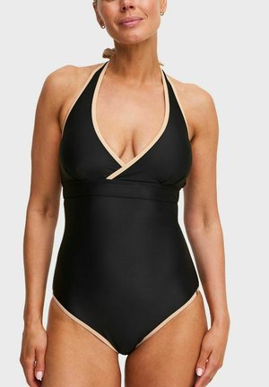 Vrouw die een zwart eendelig badpak met beige biezen en een halternek draagt, staand tegen een effen lichte achtergrond.