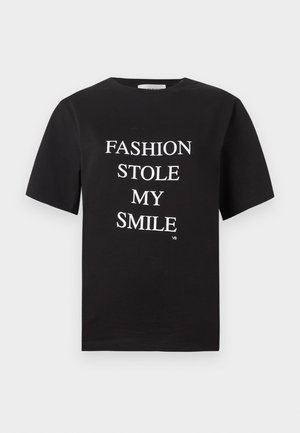 Vrouw met donker haar die een zwart T-shirt draagt met de tekst "FASHION STOLE MY SMILE", witte brede pijpen broek, zwarte sandalen met hak en tatoeages op haar armen.