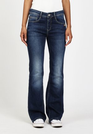 Frau trägt dunkelblaue Schlagjeans mit leichtem Verblassen und weißen Turnschuhen, steht vor einem einfarbigen Hintergrund.