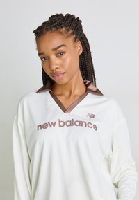 Weißes langärmliges Sportoberteil mit braunem Kragen und "New Balance"-Logo, aus atmungsaktivem Mesh-Material. Verfügt über ein V-Ausschnitt-Design.