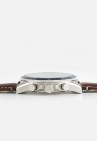 Reloj de acero inoxidable con un dial negro, acentos plateados y una correa de cuero marrón. Cuenta con dos pulsadores y un bisel texturizado.