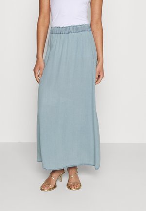 Femme portant une jupe longueur cheville bleu clair avec une taille élastique et des sandales à talons hauts dorées à lanières, se tenant devant un fond uni.