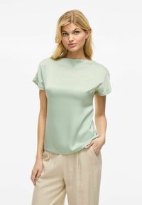 Blusa verde menta a maniche corte con una consistenza liscia e setosa e una vestibilità leggermente ampia. Abbinata a pantaloni beige chiaro plissettati.