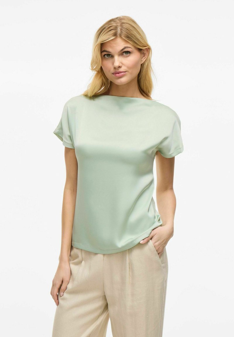 Blusa verde menta a maniche corte con una consistenza liscia e setosa e una vestibilità leggermente ampia. Abbinata a pantaloni beige chiaro plissettati.