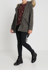 Giacca parka verde con cappuccio foderato in pelliccia, abbinata a un top floreale e jeans skinny neri. Gli stivali neri con lacci alla caviglia completano l'outfit.