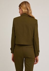 Blazer accorciato verde oliva con spalle strutturate, polsini con bottoni e una texture di tessuto liscia. La vista posteriore mette in risalto il taglio sartoriale e le linee pulite.