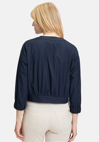 Veste cropped bleu marine en tissu lisse et léger avec des manches trois-quarts et des poignets élastiques, présentant un design simple.
