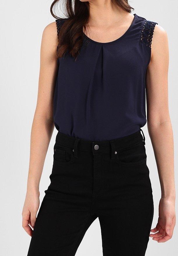 Femme portant un chemisier bleu marine sans manches avec des détails en dentelle aux épaules et un pantalon noir taille haute sur un fond uni.