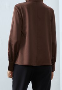 Blusa leggera marrone con maniche lunghe, polsini con bottoni e dettagli sul retro. Tessuto morbido; design su misura. Abbinata a pantaloni neri.
