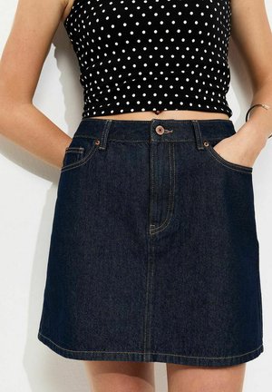 Vrouw draagt een donkerblauwe denimrok en een zwart cropped top met witte stippen, handen in de zakken van de rok, staand tegen een witte muur.