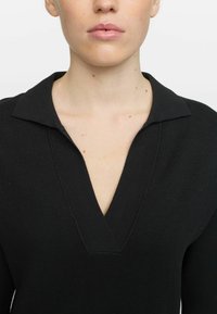 Svart stickad topp med vid V-ringning och krage. Texturerat tyg, figurnära siluett och långa ärmar. Enkel och minimalistisk design.