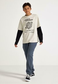 Lichtbeige grafische t-shirt met zwarte accenten, met een wereldbolontwerp en tekst. Gecombineerd met grijze joggingbroek en zwart-witte sneakers.