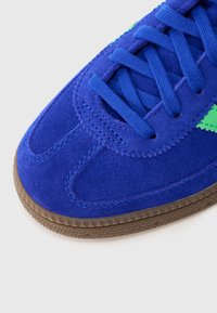 Ténis de camurça azul com atacadores azuis, detalhe de risca verde e sola de borracha texturada castanha sobre fundo branco.