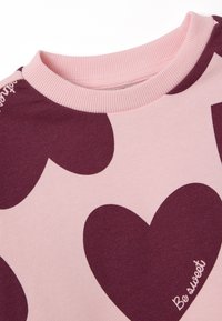 Lichtroze trui met grote donkerburgundy hartpatronen. Geribbelde halslijn en de tekst "Be sweet" geborduurd in het wit op de stof.