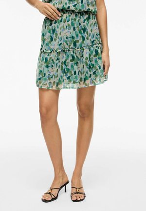 Kobieta w zielonej wzorzystej, warstwowej spódnicy i czarnych sandałach na obcasie z paskami i złotymi akcentami, stojąca na białym tle.