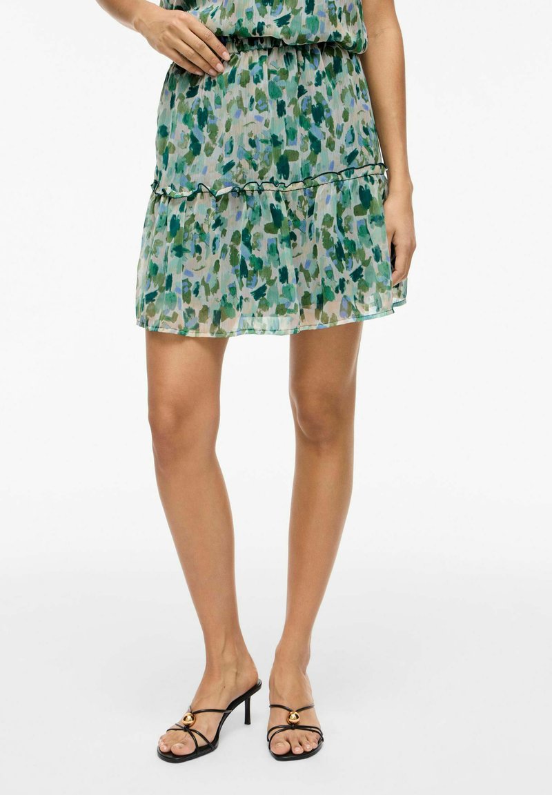 Femme portant une jupe verte à motifs superposés et des sandales à talons noires à brides avec des accents dorés, debout devant un fond blanc.