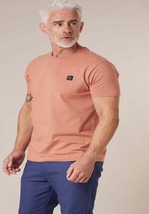 Man met wit haar en baard, gekleed in een zalmroze T-shirt en blauwe broek, kijkt naar links tegen een effen achtergrond.