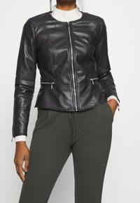 Donna che indossa una giacca di pelle nera aderente con cerniera frontale e tasche con zip, abbinata a pantaloni verde scuro e un dolcevita bianco.