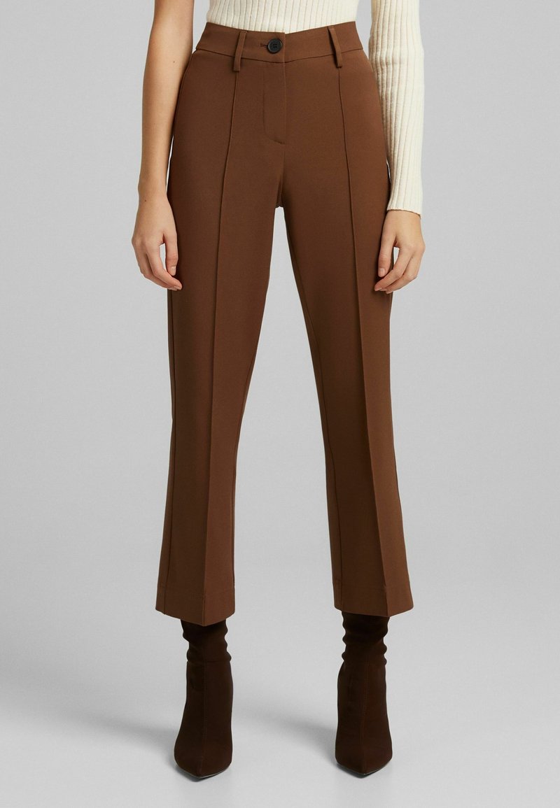 Femme portant un pantalon marron court taille haute avec des plis devant et des bottes à talons marron foncé, associé à un pull côtelé crème.