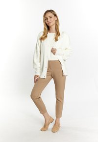 Leichte, cremefarbene Strickjacke mit Kabelmuster und Knöpfen, kombiniert mit sandfarbenen Slim-Fit-Hosen und passenden Slippern. Weiche Texturen überall.