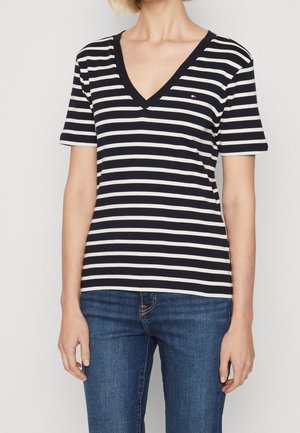 Kobieta ubrana w czarno-biały, poziomo paskowany T-shirt z krótkim rękawem i dekoltem w kształcie litery V oraz niebieskie dżinsy, stojąca na jednolitym tle.