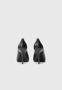 Pár černých kožen�ých stiletto podpatků zobrazených zezadu na čistém bílém pozadí.