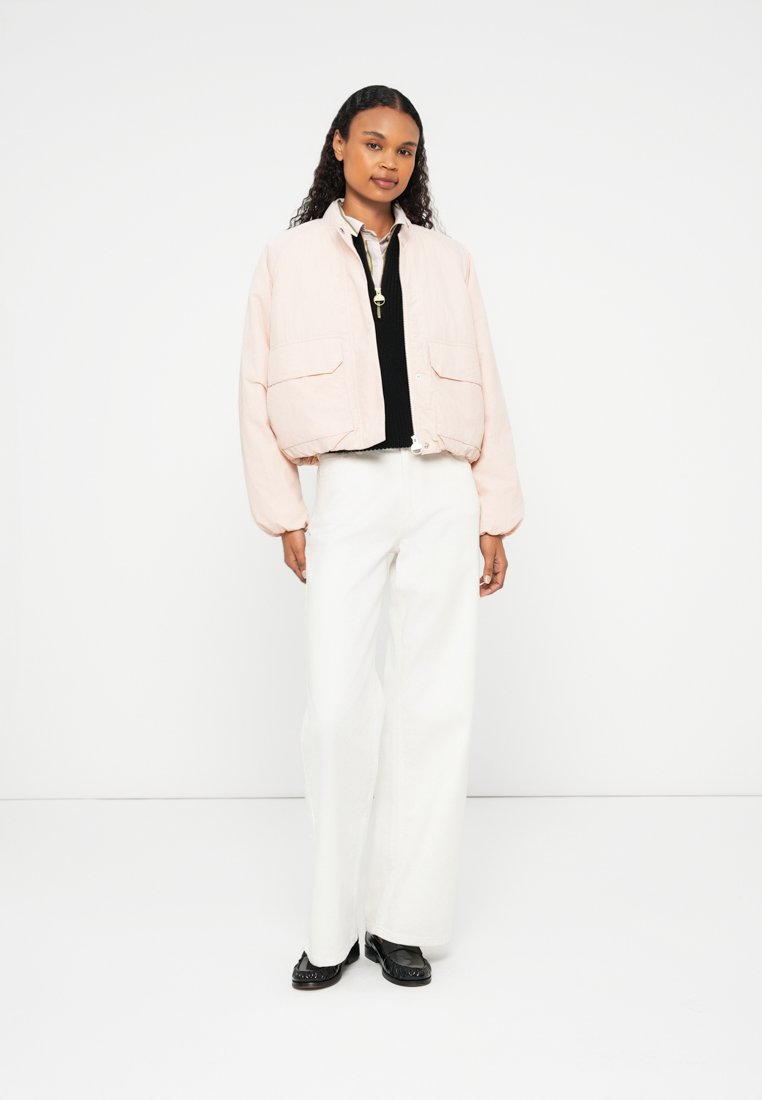 Veste bomber rose clair raccourcie avec deux poches avant, dotée d'un col rond et de poignets élastiques, associée à un pantalon large blanc.