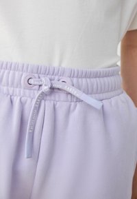 Los pantalones de chándal lila presentan una cinturilla acanalada y un cordón ajustable con herrajes circulares. Tela suave con textura mínima y detalles de costura.