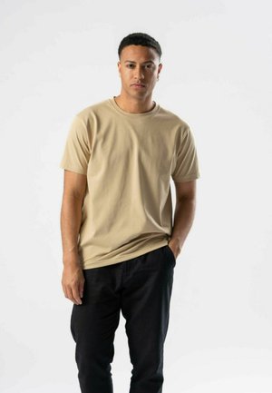 Ung mand med kort hår iført en beige t-shirt og sorte bukser, stående med den ene hånd i lommen mod en hvid baggrund.
