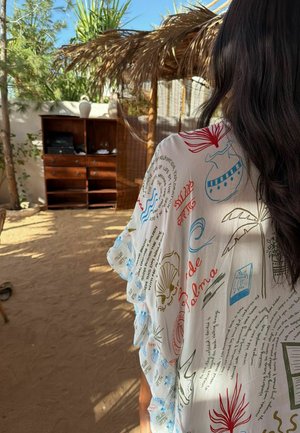 Kobieta z długimi ciemnymi włosami, ubraną w biały, wzorzysty narzutę, stojąca na nasłonecznionym, zewnętrznym terenie z piaskiem i drewnianymi meblami.