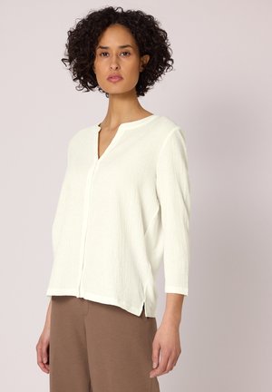 Femme aux cheveux bouclés portant un chemisier blanc à manches longues texturé et un pantalon marron, debout devant un fond clair uni.