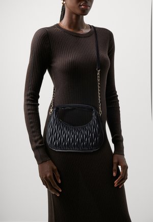 Schwarze gesteppte Handtasche mit gebogenem Griff und Kettenriemen, mit strukturierter Oberfläche und elegantem Design, getragen zu einem braunen, gerippten Kleid.