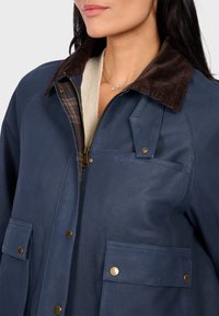 Námořnická modrá bunda s kordurovým límcem, zlatými detaily, zipovým zapínáním a dvěma předními kapsami. Vnitřní podšívka má kostkovaný vzor.