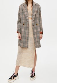 Rutig oversize blazer i grått och orange, parat med en beige stickad klänning med en sidoklyfta, och svarta sneakers med färgglada detaljer.