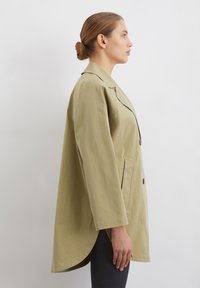Palton oversized, de culoare verde deschis, cu guler cu rever, două butoane și buzunare laterale. Prezintă o tivă curbată și mâneci lungi. Textură de material neted.