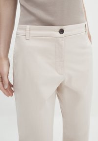 Beige katoenen broeken met een rechte snit, voorzien van een voorzak, riemlussen en een donkere knoop bij de tailleband.