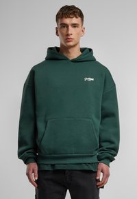 Sudadera con capucha de color verde oscuro, hecha de una tela suave, que cuenta con un bolsillo delantero, puños acanalados y un pequeño logotipo blanco en el pecho.