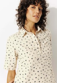 Femme aux cheveux bouclés et foncés portant une chemise crème avec de petits motifs floraux noirs et des boutons snaps, posant sur fond blanc.