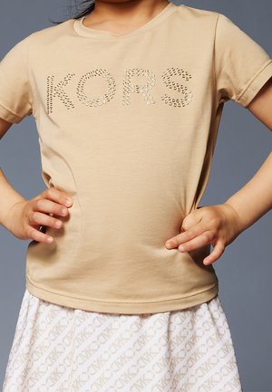 Beige Baumwoll-T-Shirt mit Rundhalsausschnitt, das mit metallic Nägeln "KORS" präsentiert. Kombiniert mit einem weißen Rock, der ein dezentes Logomuster zeigt.