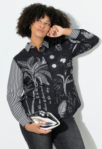 Sort skjorte med knapper, der har en blanding af palmeblade og solillustrationer samt hvid skrift på den ene side, kombineret med stribede sorte og hvide ærmer.