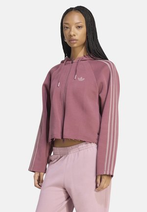 Donna con i capelli lunghi e intrecciati che indossa una felpa corta color malva con zip e strisce sulle maniche e pantaloni della tuta rosa chiaro, in piedi davanti a uno sfondo bianco.
