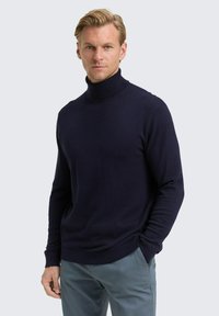 Navyblauer Rollkragenpullover in taillierter Form, aus strukturiertem Strickmaterial, mit langen Ärmeln und geripptem Saum. Kombiniert mit grauen Hosen.