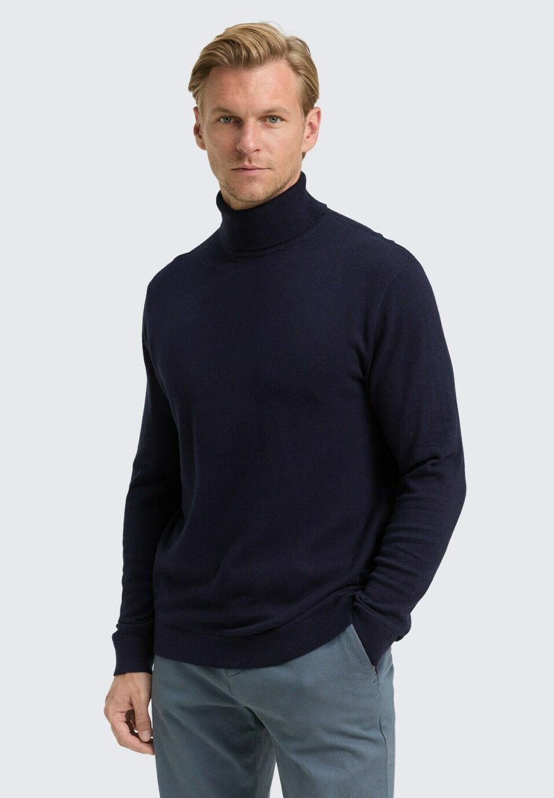 Navyblauer Rollkragenpullover in taillierter Form, aus strukturiertem Strickmaterial, mit langen Ärmeln und geripptem Saum. Kombiniert mit grauen Hosen.