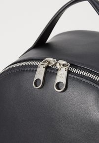 Svart läderryggsäck med silverfärgade dragkedjor och rund form. Har ett kort handtag och texturerad ytdetaljering.