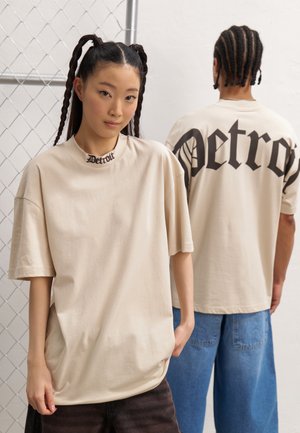 T-shirt beige oversize con stampa nera "Detroit" al collo e sul retro, con maniche corte e una texture di cotone liscia.