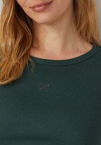 Dunkelgrünes, geripptes T-Shirt mit Rundhalsausschnitt und dezentem Logo-Detail, das auf der Brust gestickt ist. Weicher, strukturierter Stoff.